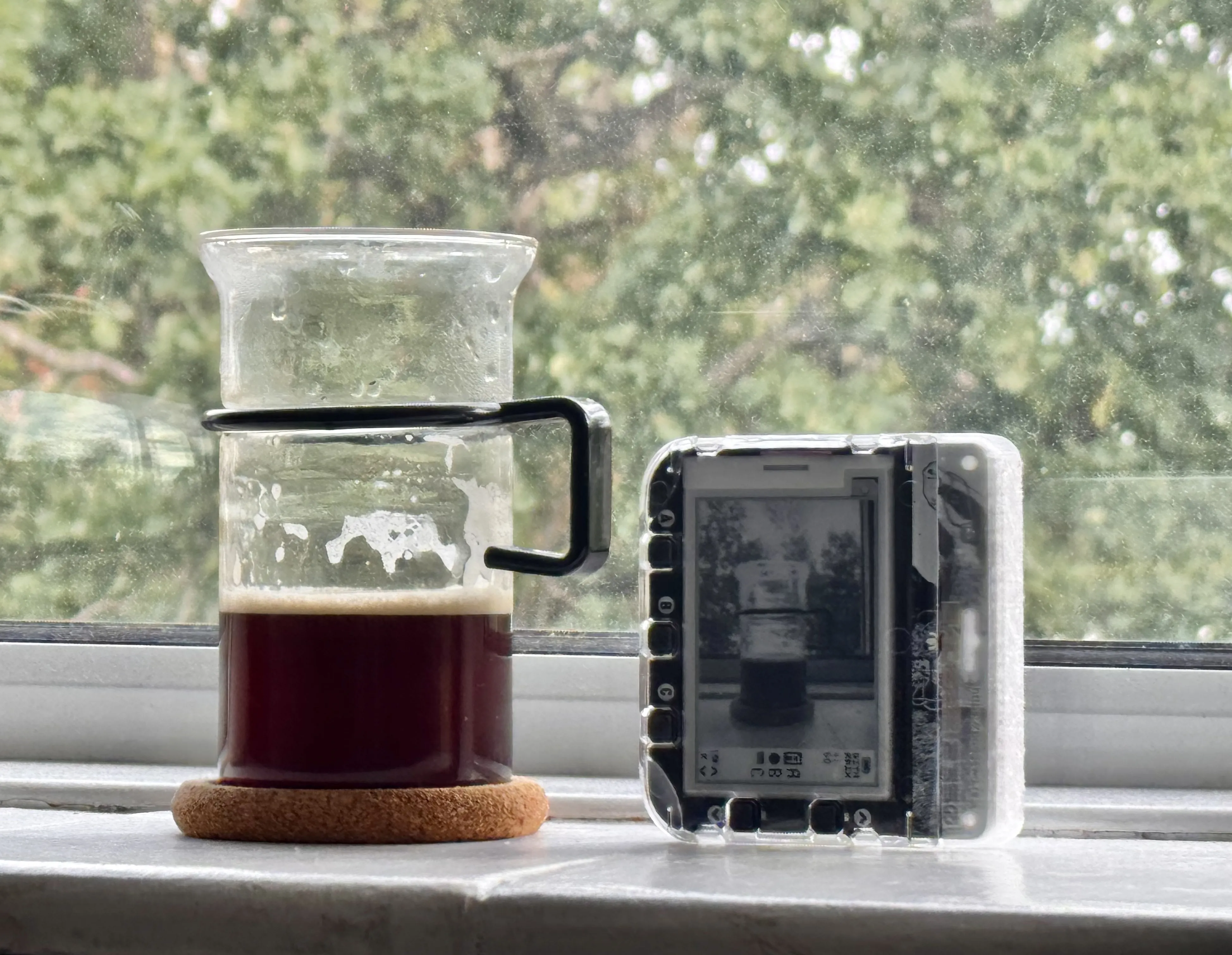 A glass coffee jug on a windowsill beside the DITHRPIX camera, its e-ink screen showing the camera's own dithered photograph of that same jug